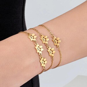 Puede incluir: Pulseras personalizadas doradas con dijes de huellas de pata. Cada dije tiene un nombre grabado. Las pulseras se usan en la muñeca. Los nombres en los dijes son Daisy, Bella, Luna, Coco, Cooper y Lucy.
