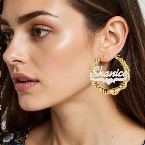 Könnte beinhalten: Goldfarbene Bambus-Creolen mit dem Namen "Shanice" in weißer Schrift. Die Ohrringe haben ein Herzdesign und sind ein stilvolles Accessoire.