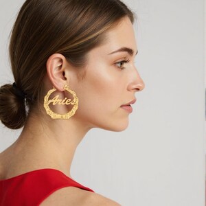 Könnte beinhalten: Goldfarbene Creolen mit dem Wort "Aries" in Schreibschrift. Die Ohrringe haben eine bambusartige Textur und werden am Ohr einer Person getragen. Die Person trägt ein rotes Oberteil.
