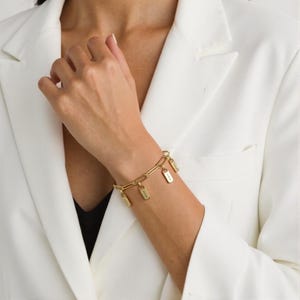 Könnte beinhalten: Goldfarbenes Gliederarmband mit mehreren rechteckigen Anhängern. Die Anhänger hängen an den Gliedern. Das Armband wird am Handgelenk getragen, vor einem weißen Blazer.