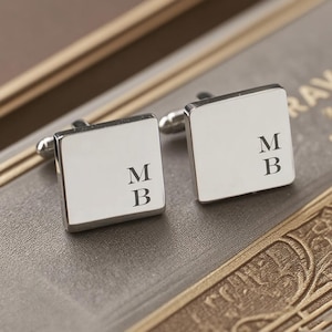 Puede incluir: Un par de gemelos cuadrados plateados, cada uno grabado con las iniciales "MB" en una fuente clásica. Los gemelos se muestran sobre una superficie texturizada de tonos neutros, sugiriendo una caja de presentación.