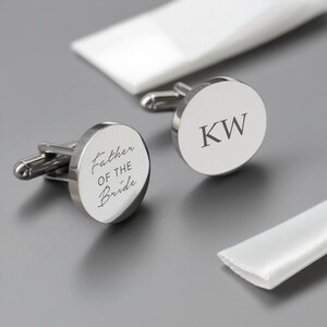 Könnte beinhalten: Silberne Manschettenknöpfe mit runder Oberfläche. Einer ist mit "Father of the Bride" graviert, der andere mit den Initialen "KW". Die Manschettenknöpfe werden mit weißer Verpackung präsentiert.