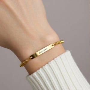 Könnte beinhalten: Ein goldfarbenes Armband mit einem rechteckigen Namensschild und einer Panzerkette. Auf dem Namensschild steht "ahive niine". Das Armband wird am Handgelenk getragen, kombiniert mit einem weißen Pullover.
