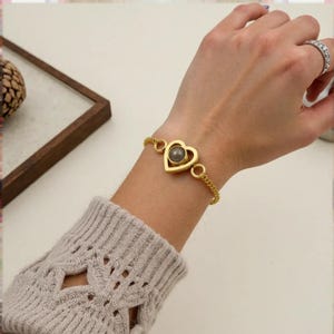 Könnte beinhalten: Goldfarbenes Armband mit herzförmigem Anhänger und grauem Stein. Das Armband wird am Handgelenk getragen, wodurch die Kette und das Design zur Geltung kommen. Der Herzanhänger ist etwa 2,5 cm breit. Ein stilvolles Accessoire.