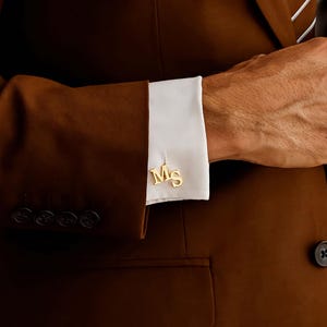 Könnte beinhalten: Nahaufnahme von Manschettenknöpfen in Gold mit den Initialen „Ms“ auf einem weißen Hemdärmel. Der Ärmel ist Teil eines braunen Anzugs. Das Bild zeigt die Details der Manschettenknöpfe und des Anzugs.