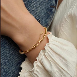 Könnte beinhalten: Ein goldenes Kettenarmband mit einem personalisierten Namensschild-Anhänger. Das Namensschild ist mit den Buchstaben "KI" graviert.