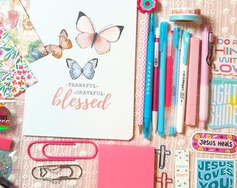 May include: A collection of stationery items including a notebook with butterfly illustrations and the word "blessed", pens, stickers, paper clips, and sticky notes. The color palette includes pink, blue, and floral patterns.