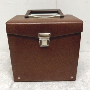 May include: A square-shaped brown storage case with a textured faux leather finish. The case features a dark brown plastic handle with a silver-toned metal accent, a central silver-toned metal latch closure, and small silver-toned metal rivets.