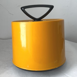 May include: A cylindrical container with a glossy mustard yellow finish and a black base. The lid features a prominent black triangular handle centered on top, providing a distinct contrast to the smooth, solid yellow body of the storage vessel.