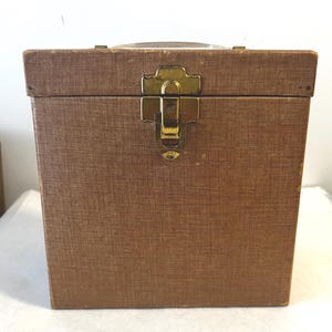 May include: A vintage brown storage case with a textured surface and a gold-colored clasp. The case has a handle on top and is rectangular in shape. The clasp is centered on the front of the case.