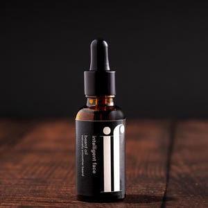 Könnte beinhalten: Eine braune Glasflasche Bartöl mit einer schwarzen Pipette. Das Etikett trägt die Aufschrift "intelligent face" und "beard oil naturally smoother beard" in weißer Schrift. Die Flasche steht auf einer Holzoberfläche.