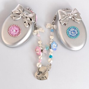 Peut inclure: Deux miroirs de poche ovales argentés avec un nœud métallique sur le dessus. Un miroir porte un emblème Hello Kitty rose avec des strass, l'autre un bleu. Une breloque perlée avec des fleurs et un cœur portant l'inscription BEST FRIENDS.