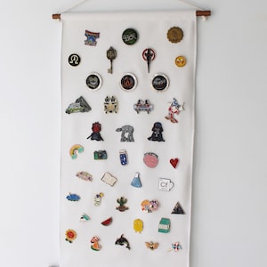 May include: A white rectangular fabric wall banner suspended from a wooden dowel with a cord. The banner displays a collection of various colorful enamel pins arranged in rows, featuring diverse shapes including symbols, characters, and objects.