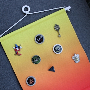 Pode incluir: Uma faixa decorativa com um gradiente de amarelo a laranja, exibindo vários pinos e uma chave. Os pinos incluem um personagem de desenho animado, uma chave e designs circulares com texto. A faixa tem uma haste e um cordão brancos.