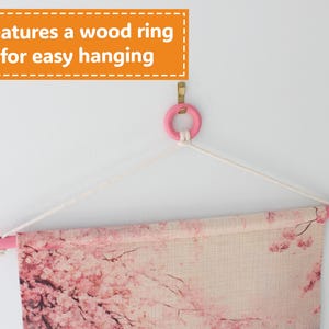 May include: A wall hanging featuring a pink wooden rod and ring, with a floral design. The text "Features a wood ring for easy hanging" is displayed in an orange box. The artwork is suspended by a white rope.