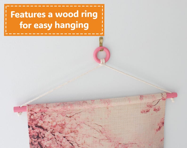May include: A wall hanging featuring a pink wooden rod and ring, with a floral design. The text "Features a wood ring for easy hanging" is displayed in an orange box. The artwork is suspended by a white rope.