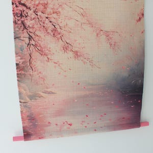 May include: A wall hanging featuring a serene landscape with cherry blossoms. The artwork depicts a pink and white floral scene with a body of water and falling petals. The bottom has a pink dowel for hanging.