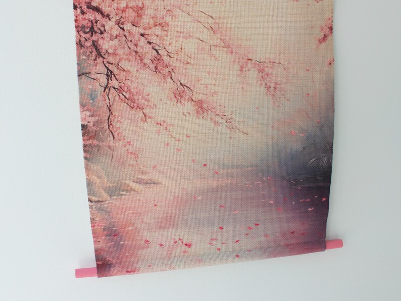 May include: A wall hanging featuring a serene landscape with cherry blossoms. The artwork depicts a pink and white floral scene with a body of water and falling petals. The bottom has a pink dowel for hanging.