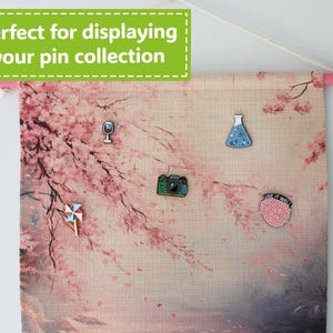 May include: A fabric banner with a pink wooden rod at the top, displaying a cherry blossom design and several enamel pins. The pins include a wine glass, a camera, a pinwheel, a science beaker, and a brain with the text "USE IT WELL". The banner has the text "Perfect for displaying your pin collection".
