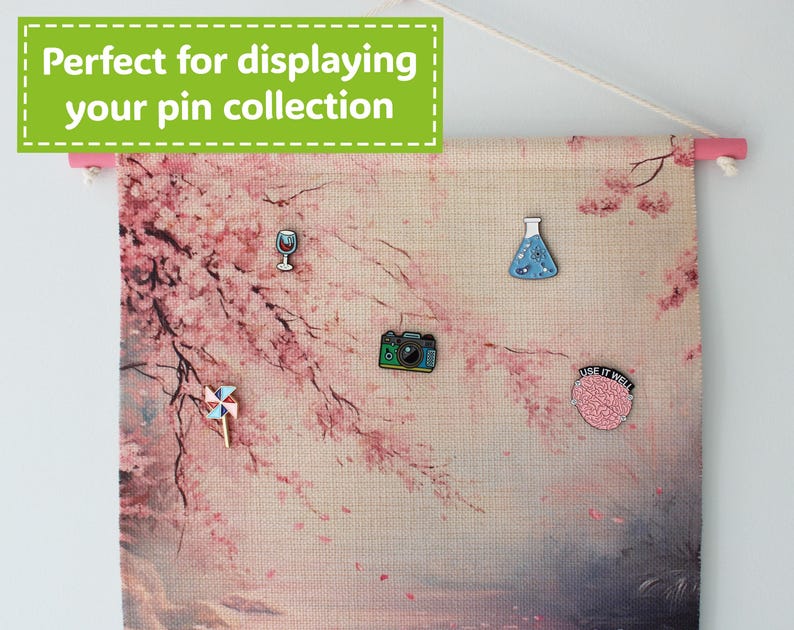 May include: A fabric banner with a pink wooden rod at the top, displaying a cherry blossom design and several enamel pins. The pins include a wine glass, a camera, a pinwheel, a science beaker, and a brain with the text "USE IT WELL". The banner has the text "Perfect for displaying your pin collection".
