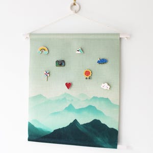 Könnte beinhalten: Eine Wandbehänge mit einem Berglandschaftsdesign in Grün und Blau. Der Stoffbanner hat oben und unten eine Holzleiste. Dekorative Anstecknadeln mit Regenbogen, Kamera und anderen Formen sind am Banner befestigt.