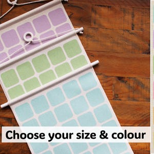 May include: Wall hanging featuring a repeating square design in pastel shades of lavender, green, and blue. The text "Choose your size & colour" is visible. The hanging is suspended by a white cord and wooden dowels, with a wooden background.