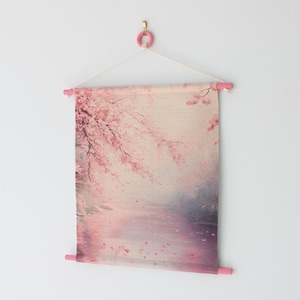 May include: A wall hanging featuring a serene landscape with pink cherry blossoms. The artwork is displayed on a light-colored fabric, held by pink wooden dowels and a pink hook. The scene depicts a river and falling petals.