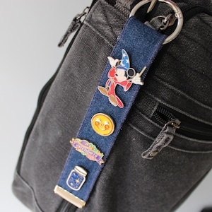 Può includere: Una striscia di tessuto denim blu scuro fissata a un anello di metallo argentato, con quattro spille smaltate: un topo mago in tunica rossa, un sole giallo sorridente, uno stendardo viola con il testo OVERTHINKER e un vasetto blu con stelle.