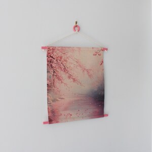 May include: A fabric wall hanging featuring a pink and white cherry blossom design. The artwork is suspended from a pink wooden rod and a white string, creating a minimalist aesthetic. The image is mounted on a white wall.