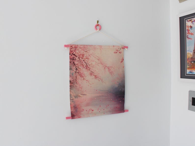 May include: A fabric wall hanging featuring a pink and white cherry blossom design. The artwork is suspended from a pink wooden rod and a white string, creating a minimalist aesthetic. The image is mounted on a white wall.