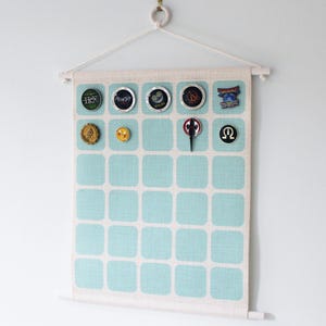 May include: A fabric display board with a light blue and white square pattern, designed for displaying pins and badges. The board has a wooden rod at the top and is decorated with various pins and buttons. The board is suspended by a white rope.