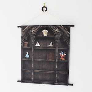 Könnte beinhalten: Dunkelbrauner Wandbehang mit einem Druck einer gotischen Bücherregal-Darstellung. Die Regale zeigen Bücher, eine Flasche und eine Mickey Mouse-Figur. Der Behang wird von einer schwarzen Stange und einer weißen Schnur gehalten.