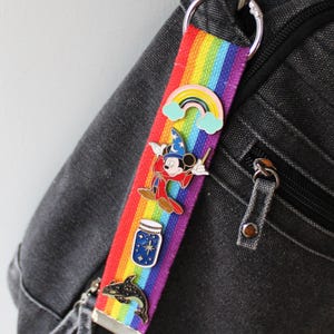May include: A rainbow-striped lanyard with a silver keyring attached to a black backpack. The lanyard features enamel pins of a rainbow, Mickey Mouse as a wizard, a jar of stars, and a dolphin.