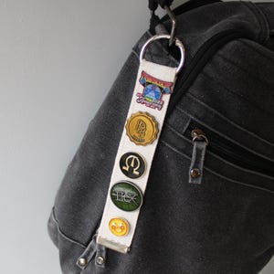 May include: A white fabric keychain with several decorative pins attached to a black backpack. The pins are round and feature various designs and colours. The keychain is attached to a silver metal ring.