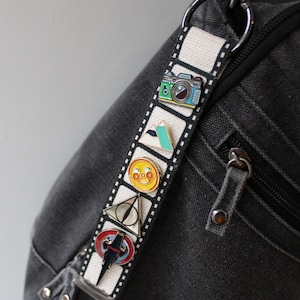 May include: A black denim bag with a white strap featuring colorful enamel pins. The pins include a camera, mountain, smiley face, triangle, and a lightning bolt design. The bag has a zipper and silver hardware.