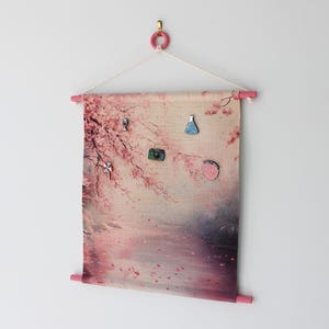 May include: A fabric wall hanging with a pink and beige floral design, featuring a pink wooden dowel at the top and bottom. Several decorative pins are attached to the fabric. The hanging is suspended from a pink ring.
