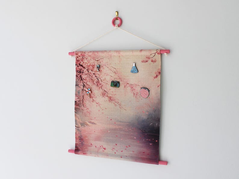 May include: A fabric wall hanging with a pink and beige floral design, featuring a pink wooden dowel at the top and bottom. Several decorative pins are attached to the fabric. The hanging is suspended from a pink ring.