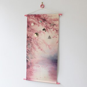 May include: A rectangular wall hanging with a pink floral design, featuring cherry blossoms and a serene landscape. The fabric is held by pink wooden dowels and a white hanger. Several decorative magnets are attached.
