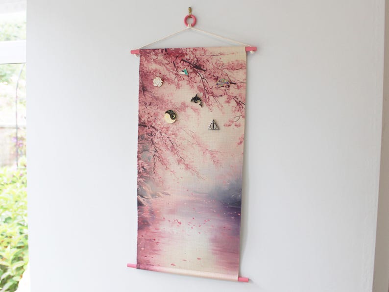 May include: A rectangular wall hanging with a pink floral design, featuring cherry blossoms and a serene landscape. The fabric is held by pink wooden dowels and a white hanger. Several decorative magnets are attached.