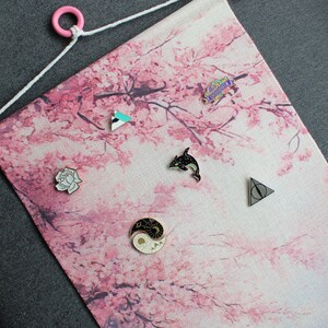May include: A pink and white fabric banner with a cherry blossom design, featuring various enamel pins. The pins include a dolphin, a yin-yang symbol, a geometric shape, a rose, and a symbol from a popular book series. The banner has a pink wooden dowel and a white string for hanging.