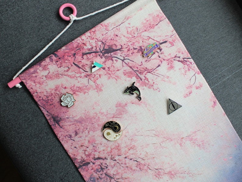 May include: A pink and white fabric banner with a cherry blossom design, featuring various enamel pins. The pins include a dolphin, a yin-yang symbol, a geometric shape, a rose, and a symbol from a popular book series. The banner has a pink wooden dowel and a white string for hanging.