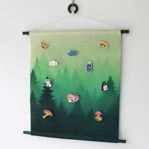May include: A tapestry featuring a forest scene in shades of green, decorated with enamel pins. The pins depict a rainbow, cloud, jar, cat, mushroom, and other nature-inspired designs. The tapestry is suspended from a wooden rod.