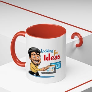 Puede incluir: Taza de cerámica blanca con borde y asa rojos. La taza presenta una ilustración de dibujos animados de un hombre con barba mirando la pantalla de un portátil. El texto "Looking for Ideas" está impreso en la taza.