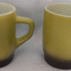 May include: Two matching mugs with a smooth, light olive green finish on the upper portion and a dark brown, ribbed base. Each mug features a sturdy, integrated handle on the side and a clean, white interior, presenting a simple, functional design.