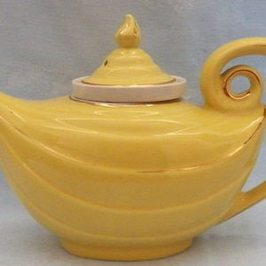 May include: A bright yellow ceramic teapot featuring a curved handle with a circular cutout, a pointed spout, and a matching lid with a finial. The teapot is decorated with subtle horizontal ridges and thin gold trim along the edges.