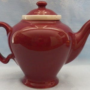 May include: A glossy deep red ceramic teapot featuring a rounded body, a curved handle, and a pointed spout. The teapot includes a matching red lid with a small knob handle, accented by a contrasting white circular rim around the lid base.