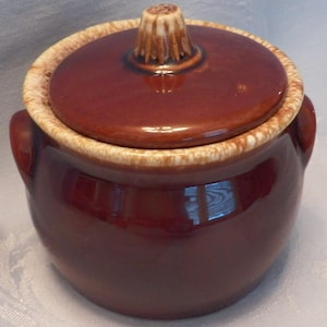May include: A round, dark brown ceramic bean pot with a matching lid. The pot features two side handles and a decorative, textured rim with a cream-colored drip glaze. The lid has a central, ridged knob handle with a similar cream-colored finish.