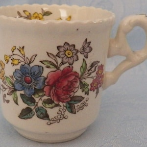 May include: Cream-colored teacup with a floral design featuring red, blue, yellow, and pink flowers with green leaves. The cup has a scalloped rim and a curved handle.