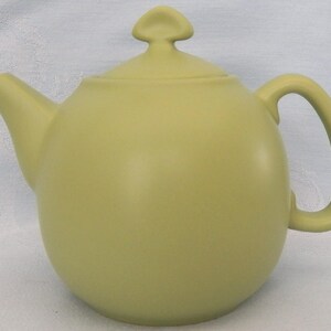 May include: A matte light olive green teapot with a rounded body, a curved spout, a C-shaped handle, and a matching lid featuring a small, triangular-shaped knob on top. The teapot has a smooth, solid-colored finish and a simple, functional design.