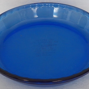 May include: A round, cobalt blue glass pie dish featuring a smooth interior surface, a fluted rim design, and two integrated side handles for gripping. The center of the dish displays embossed text that is partially visible through the glass.